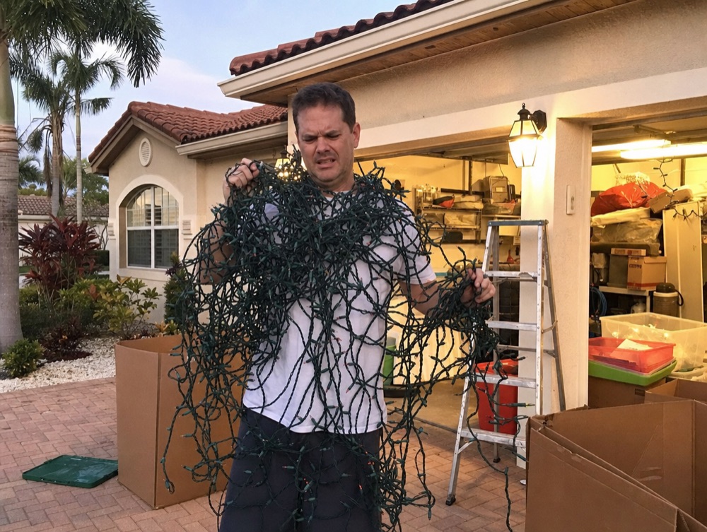 Frustrated homeowner tangled in a giant ball of Christmas lights next to a ladder and storage bins